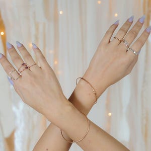 A close-up of a person's hands wearing multiple rings and bracelets against a white background with a blurred, golden-colored curtain.