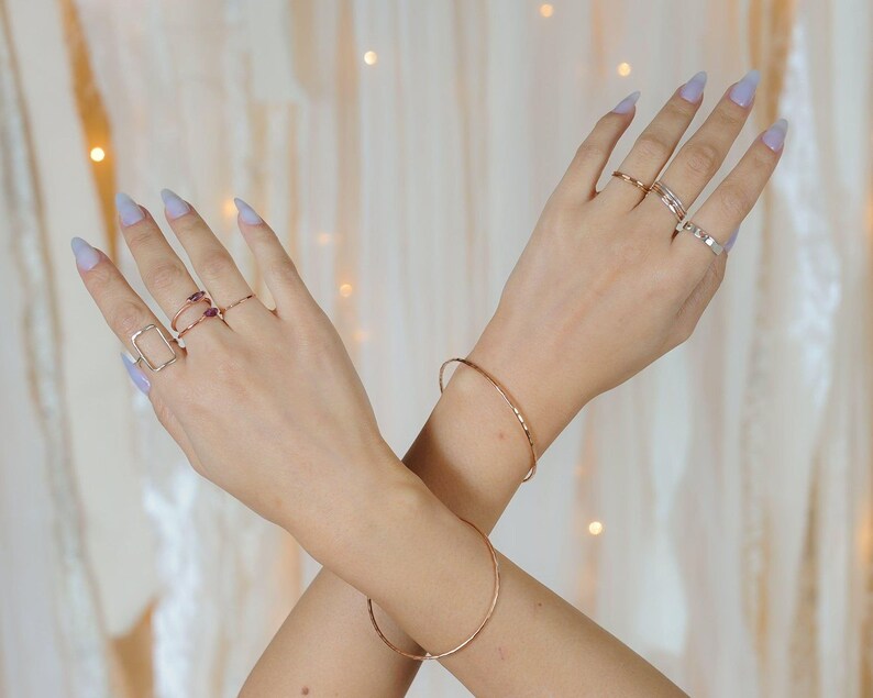 A close-up of a person's hands wearing multiple rings and bracelets against a white background with a blurred, golden-colored curtain.