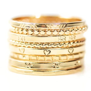 May include: A stack of seven gold rings with different engraved designs, including stars, hearts, and a clover. The rings are stacked on top of each other, creating a layered effect.