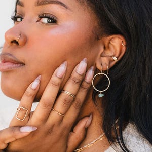A close-up of a woman's face, with her hand resting on her chin. She is wearing multiple rings and earrings, including a nose ring and hoop earrings.