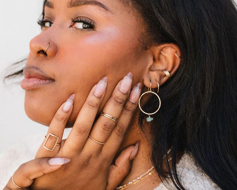 A close-up of a woman's face, with her hand resting on her chin. She is wearing multiple rings and earrings, including a nose ring and hoop earrings.