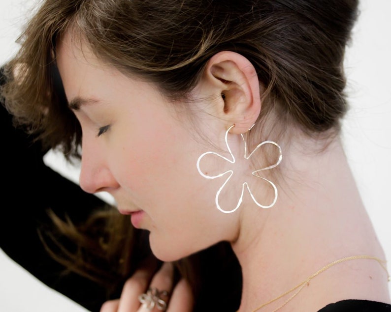 May include: Silver flower-shaped hoop earrings. The earrings are large and have a delicate, hammered texture.