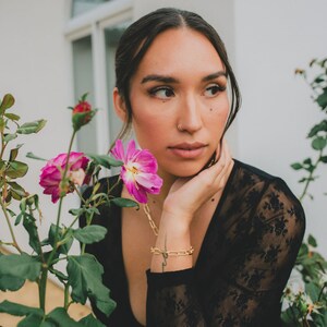 Puede incluir: Una mujer con cabello oscuro y un aro en la nariz lleva una blusa de encaje negro y mira hacia un lado. Est&aacute; sentada cerca de un arbusto de flores rosas.