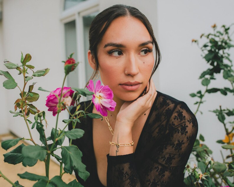Puede incluir: Una mujer con cabello oscuro y un aro en la nariz lleva una blusa de encaje negro y mira hacia un lado. Est&aacute; sentada cerca de un arbusto de flores rosas.