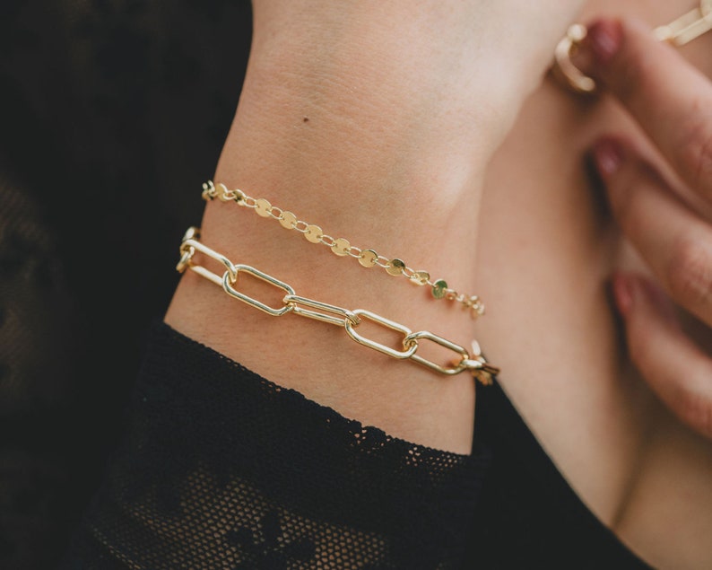 Puede incluir: Dos pulseras de oro en la mu&ntilde;eca de una persona. La pulsera superior es una cadena delicada con peque&ntilde;os eslabones redondos. La pulsera inferior es una cadena m&aacute;s gruesa con eslabones ovalados m&aacute;s grandes.