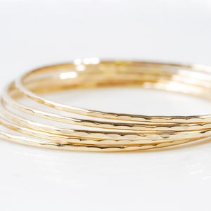 Image shows close up of our ultra thin 14 karat gold bangles. Each one is hand hammered for a light texture. Order one or more in fine 14 karat yellow gold filled. Elevate your stack. Beautiful, polished, shiny. Bracelet width is 1/16 inch wide.