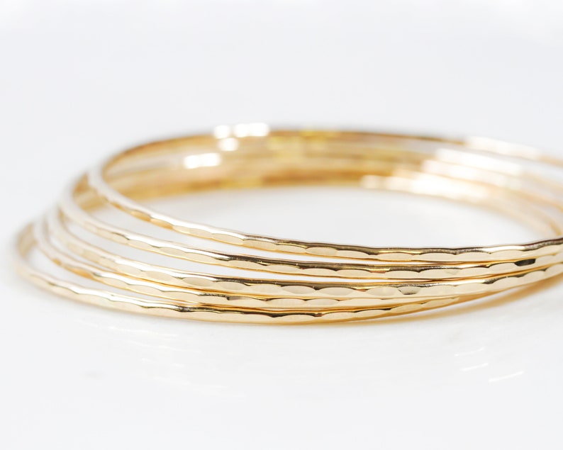 Image shows close up of our ultra thin 14 karat gold bangles. Each one is hand hammered for a light texture. Order one or more in fine 14 karat yellow gold filled. Elevate your stack. Beautiful, polished, shiny. Bracelet width is 1/16 inch wide.