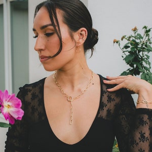 Puede incluir: Una mujer con un top de encaje negro, un collar de cadena dorada y una pulsera de cadena dorada. El collar tiene un colgante grande y redondo.