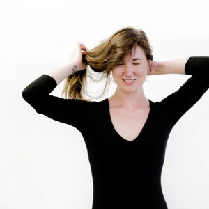 May include: A woman with long brown hair wearing a black long-sleeved shirt with a v-neck. She is smiling and has her hair pulled back with one hand.