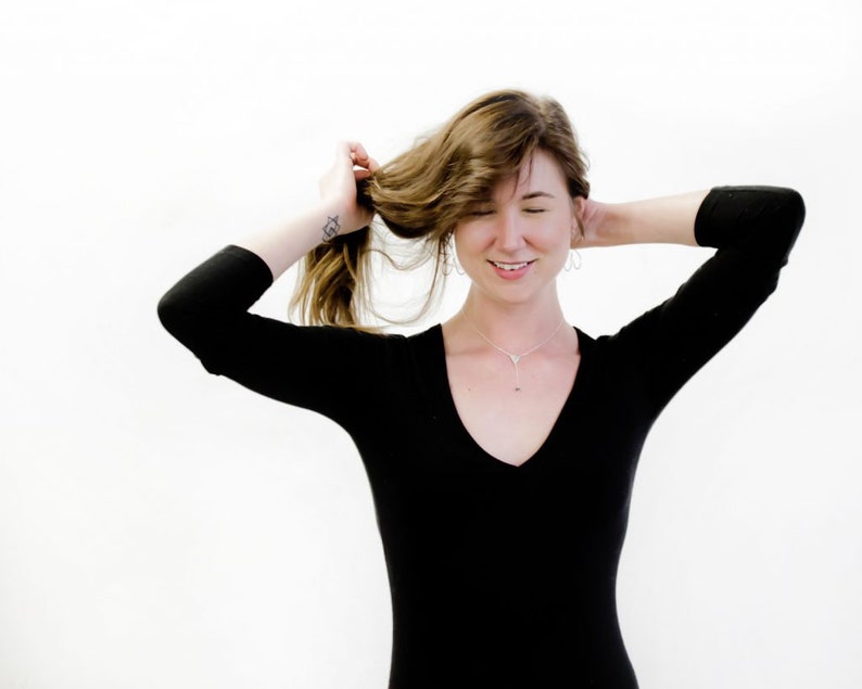 May include: A woman with long brown hair wearing a black long-sleeved shirt with a v-neck. She is smiling and has her hair pulled back with one hand.