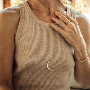 Close up shows model wearing the 14 karat yellow gold filled moon worn long at 28 inches. Each moon pendant is hand forged by our team of women artists. The crescent moon is hand hammered and the moon itself measures 44mm in length.
