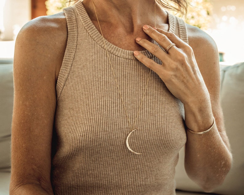 Close up shows model wearing the 14 karat yellow gold filled moon worn long at 28 inches. Each moon pendant is hand forged by our team of women artists. The crescent moon is hand hammered and the moon itself measures 44mm in length.