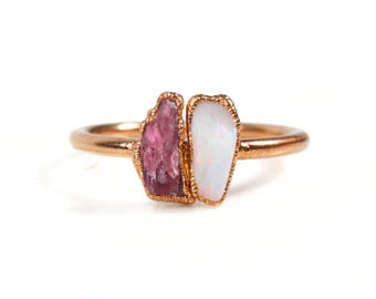 Può includere: Un anello in oro rosa con una gemma rosa grezza e un'opale bianca. Le gemme sono affiancate, creando un design unico e organico. La fascia dell'anello è in metallo lucido.
