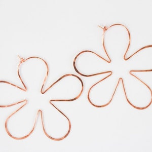 May include: A pair of large, copper-colored flower-shaped hoop earrings. The earrings are made of a single piece of wire that is bent into a flower shape.