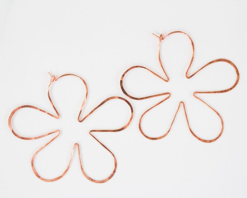 May include: A pair of large, copper-colored flower-shaped hoop earrings. The earrings are made of a single piece of wire that is bent into a flower shape.