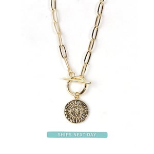 May include: A gold necklace with a toggle clasp and a circular pendant. The pendant has a sunburst design. The necklace features an elongated link chain. The text "SHIPS NEXT DAY" is at the bottom.
