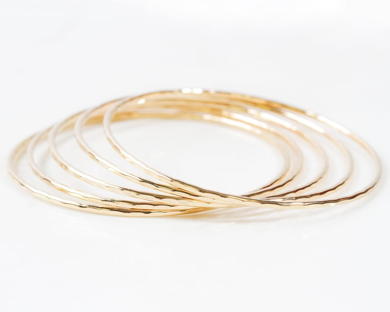 Image shows close up of the exquisite texture of our 14 karat yellow gold filled bangles. Showing five stunning bangles, each bracelet is hand hammered which gives them their special sparkle. Wear individually or stack them up to create a unique set.