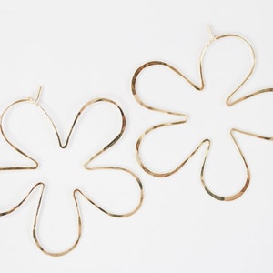 May include: A pair of gold flower-shaped hoop earrings. The earrings are made of a thin, hammered metal and have a delicate, feminine look.
