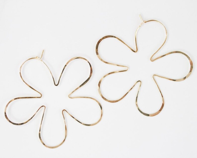 May include: A pair of gold flower-shaped hoop earrings. The earrings are made of a thin, hammered metal and have a delicate, feminine look.