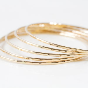 Image shows close up of the exquisite texture of our 14 karat yellow gold filled bangles. Showing five stunning bangles, each bracelet is hand hammered which gives them their special sparkle. Wear individually or stack them up to create a unique set.