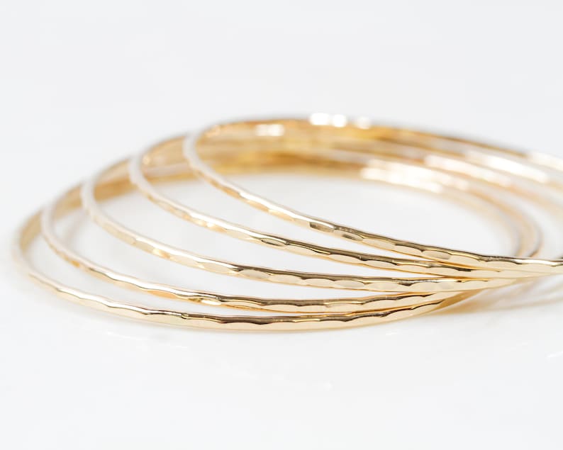 Image shows close up of the exquisite texture of our 14 karat yellow gold filled bangles. Showing five stunning bangles, each bracelet is hand hammered which gives them their special sparkle. Wear individually or stack them up to create a unique set.