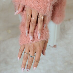 Pu&ograve; includere: Primo piano di mani con diversi anelli d'oro su diverse dita. Le unghie sono dipinte con uno smalto glitter bianco.