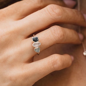 Può includere: Un anello dorato con tre pietre grezze di forma irregolare, nere, trasparenti e azzurre. L'anello è indossato su una mano, con una delicata collana visibile sullo sfondo. Le pietre sono incastonate in uno stile minimalista.