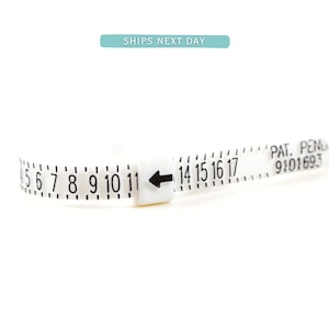 Puede incluir: Un medidor de talla de anillo de plástico blanco con marcas negras. Los números del 5 al 17 son visibles. La flecha apunta al número 14. El texto "PAT. PEND. 9101693" está impreso en el medidor.