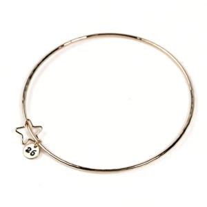 May include: A silver-colored bangle bracelet with a star charm and a small round tag with the number 26. The bracelet is circular and has a hammered texture. The star charm is hollow and has five points.