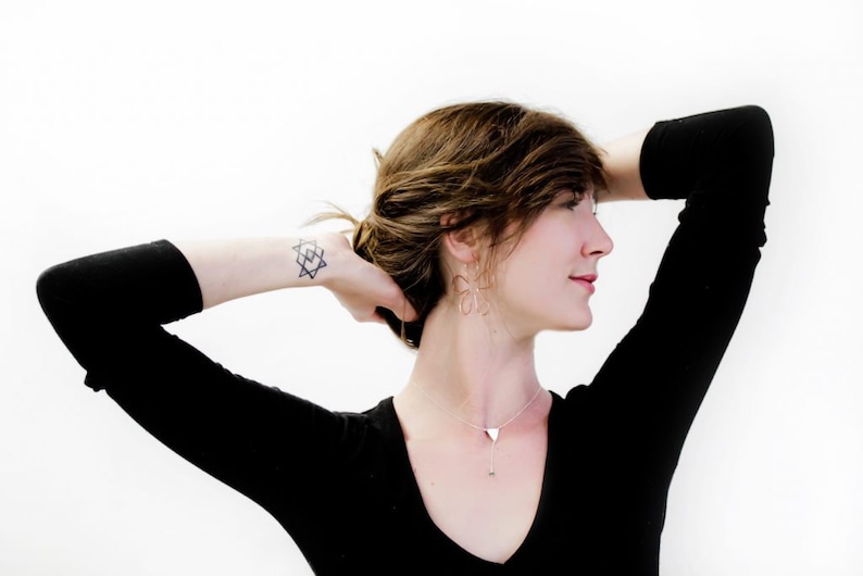 May include: A woman with brown hair wearing a black long-sleeved shirt and silver jewelry. She has a geometric tattoo on her wrist.