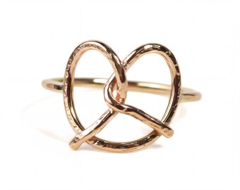 May include: A gold-toned ring featuring a central pretzel-shaped design. The metal surface of the pretzel motif has a hammered, textured finish, while the circular band of the ring remains smooth and polished, creating a distinct contrast.