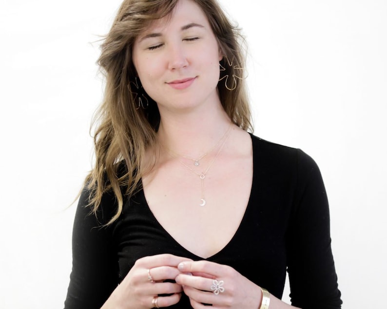 May include: A woman wearing a black v-neck shirt with three gold necklaces, one with a crescent moon pendant. She is wearing a gold ring on her left hand and a silver ring on her right hand. She is also wearing a silver ring on her left ring finger and a silver ring on her right ring finger.