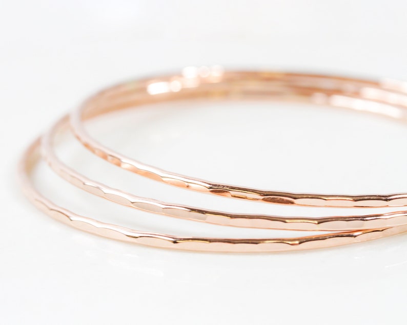 Image shows close up of our ultra thin 14 karat rose gold bangles. Hand hammered for a light texture, each bracelet is high quality and made to stand the test of time.  At a width of 1/16 inch, each stacking bangle will glint and glisten in the sun.