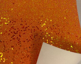 May include: Orange glitter fabric with a white backing. The fabric is covered in small, sparkling glitter particles in shades of orange, yellow, and red. The white backing is visible where the fabric is folded.