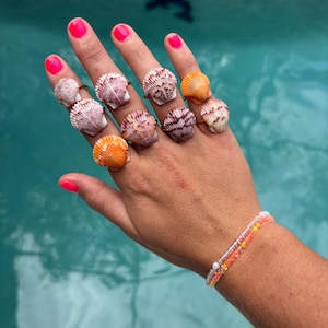 Puede incluir: Una mano con múltiples anillos de conchas marinas en varios colores, incluyendo rosa, naranja y blanco. Los anillos están engarzados en bandas doradas. También se ve una pulsera de cuentas. El fondo es una piscina turquesa.