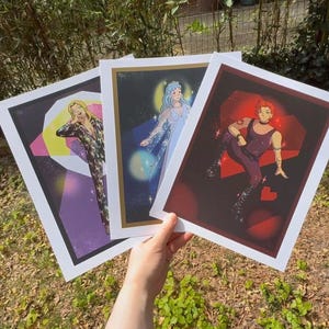 May include: Three art prints featuring stylized illustrations. One print showcases a figure in a patterned jumpsuit, another depicts a figure with blue hair and a flowing gown, and the third features a figure in a dark outfit with red accents. Each print has a white border.