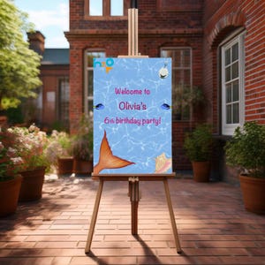 May include: A wooden easel displays a party sign with a blue background and the text "Welcome to Olivia's 6th birthday party!". The sign features a mermaid tail, a seashell, and two fish illustrations. The sign is outdoors.