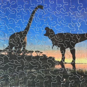 May include: A completed jigsaw puzzle featuring a prehistoric scene with two dinosaur silhouettes against a blue sky and sunset backdrop. The puzzle pieces are visible, creating a textured effect. The dinosaurs are a long-necked Brachiosaurus and a Tyrannosaurus Rex.