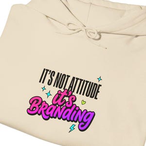 May include: A cream-colored hooded sweatshirt featuring the black text IT'S NOT ATTITUDE and the purple text it's BRANDING. The design includes small cyan four-pointed stars, a small green heart, and a small cyan lightning bolt graphic.