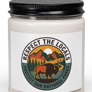 May include: A white candle jar with a black lid. The label features a circular design with the text "Respect the Locals" and "Protect Our National Parks." The design includes a bison, mountains, trees, and a sun.