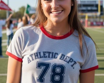 May include: A gray ringer t-shirt with red trim and the text "ATHLETIC DEPT 78" in navy blue. The shirt is worn by a person smiling on a football field.