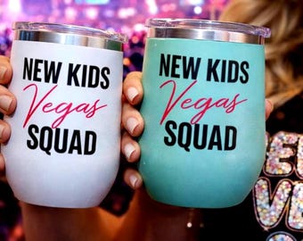 May include: Two stainless steel tumblers with lids, one white and one light teal. Both feature the black text "NEW KIDS" and "SQUAD" with the red script word "Vegas" in the center. The tumblers have a rounded shape and metallic rims.