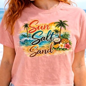 May include: A light pink crop top with the words "Sun Salt Sand" in a colorful design. The graphic includes palm trees, waves, and tropical fruits. The model is wearing blue denim shorts.