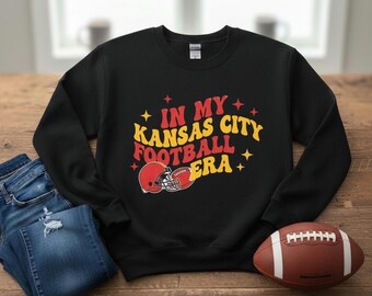 May include: Black sweatshirt with the text "IN MY Kansas City FOOTBALL ERA" in yellow and red. A red football helmet graphic is below the text. A brown football and blue jeans are also in the image.