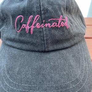 May include: A dark gray baseball cap with the word "Caffeinated" embroidered in pink script across the front. The cap has a curved brim and a classic six-panel design, suitable for casual wear.