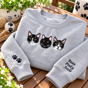 Puede incluir: Una sudadera gris claro de cuello redondo con un diseño bordado de tres caras de gato en el pecho. La manga izquierda tiene dos huellas de pata negras bordadas, y la manga derecha presenta los nombres bordados Bean, Pinky y Josie.