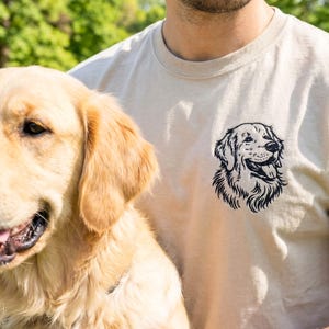 A sand-colored t-shirt laid on grass featuring a black thread embroidered Golden Retriever portrait on the left chest. The left sleeve is embroidered with the name “Bruno” and a small paw print detail.