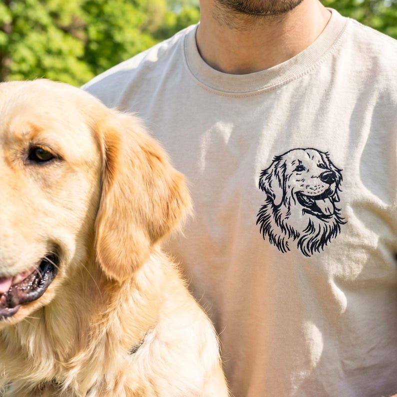 A sand-colored t-shirt laid on grass featuring a black thread embroidered Golden Retriever portrait on the left chest. The left sleeve is embroidered with the name “Bruno” and a small paw print detail.