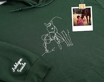 May include: Dark green hoodie with a white embroidered horse design on the front. A small photo is clipped to the hoodie. The sleeve has the embroidered text "Ashlynn Suede".