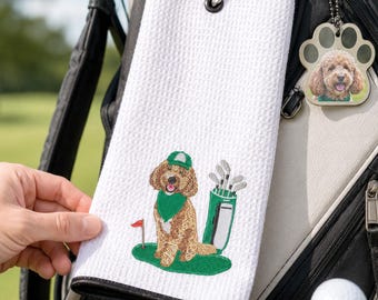 May include: White golf towel with a black border, featuring an embroidered cartoon dog wearing a green hat and bandana. A golf bag charm with a dog's face hangs nearby. A golf ball and tee are in the foreground.